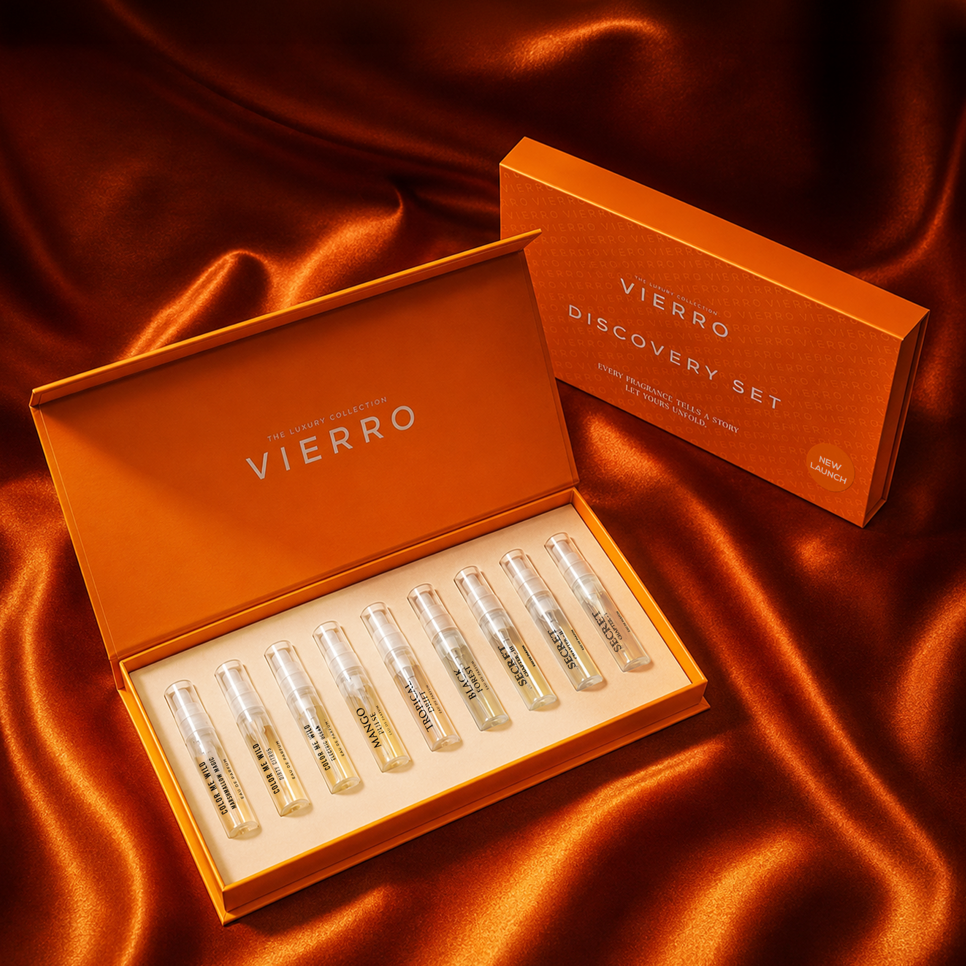 An open orange VIERRO New Arrivals Discovery Set box with eight small glass sample vials arranged in a row on a cream insert, with a matching closed VIERRO box standing behind it on draped satin fabric.